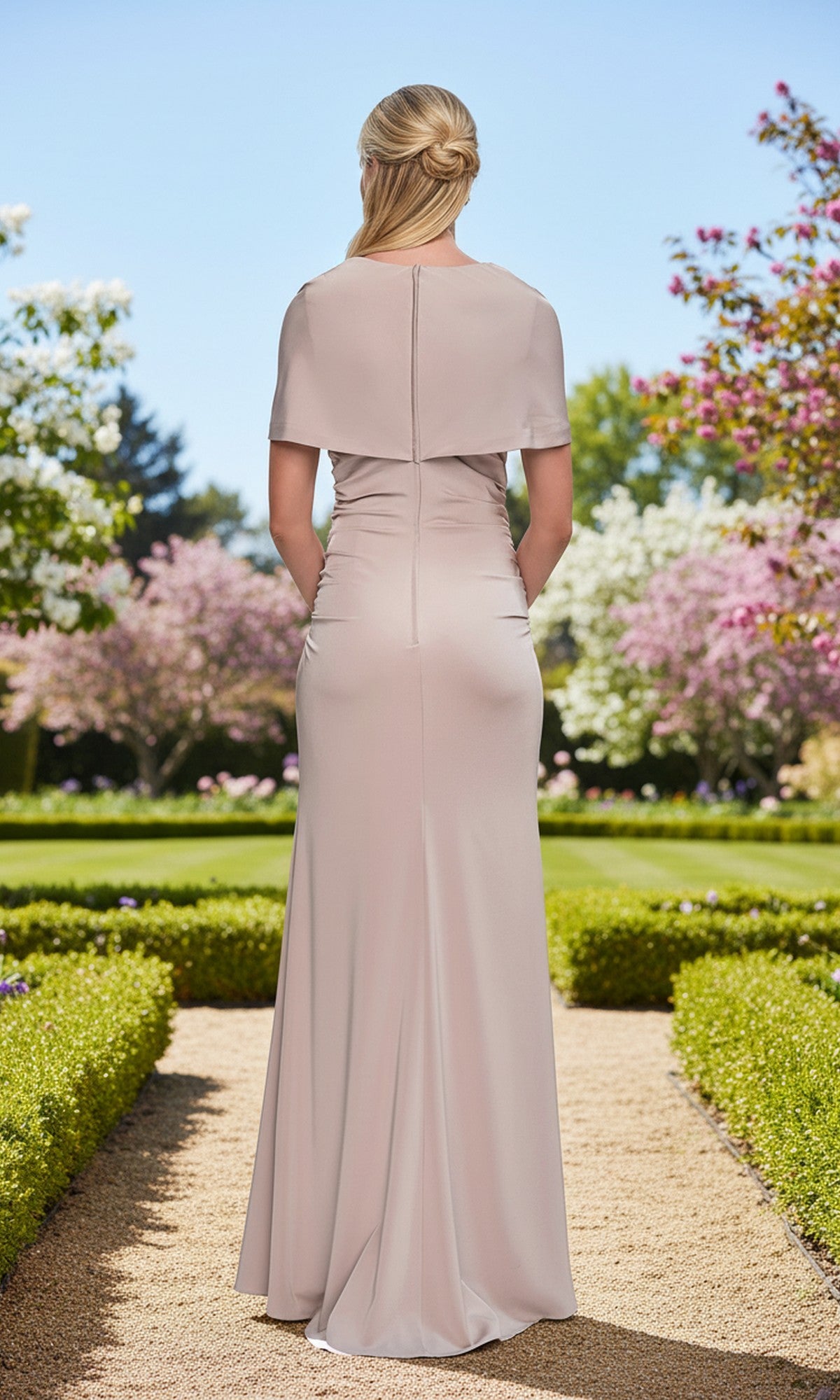 A woman stands on a garden path with her back to the camera, surrounded by blooming trees and hedges, wearing the Long Prom Dress: La Femme 33157—a pale pink formal sheath dress with a v-neckline and short sleeves.